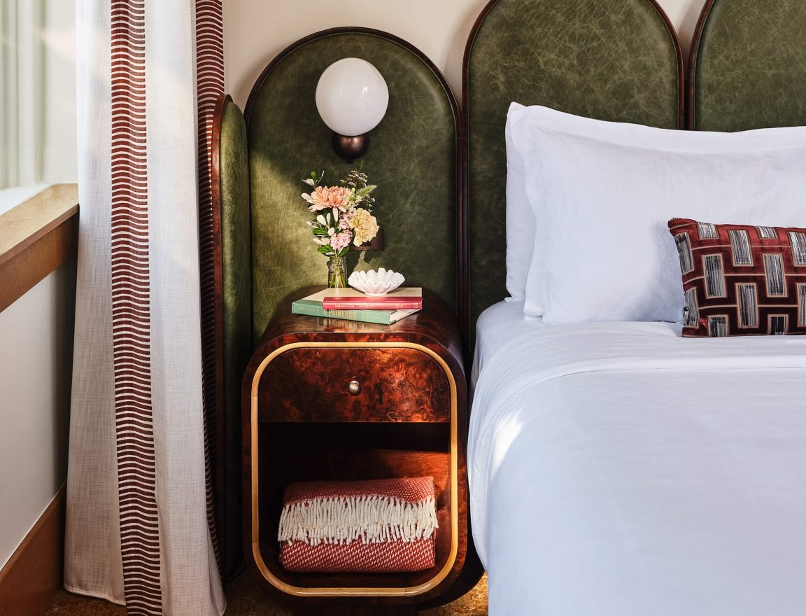 The bedside area of the Deluxe Classic Guest room at the Crawford Hotel in Denver, Colorado. The area has a small table with an extra blanket, fresh cut flowers and a lamp.