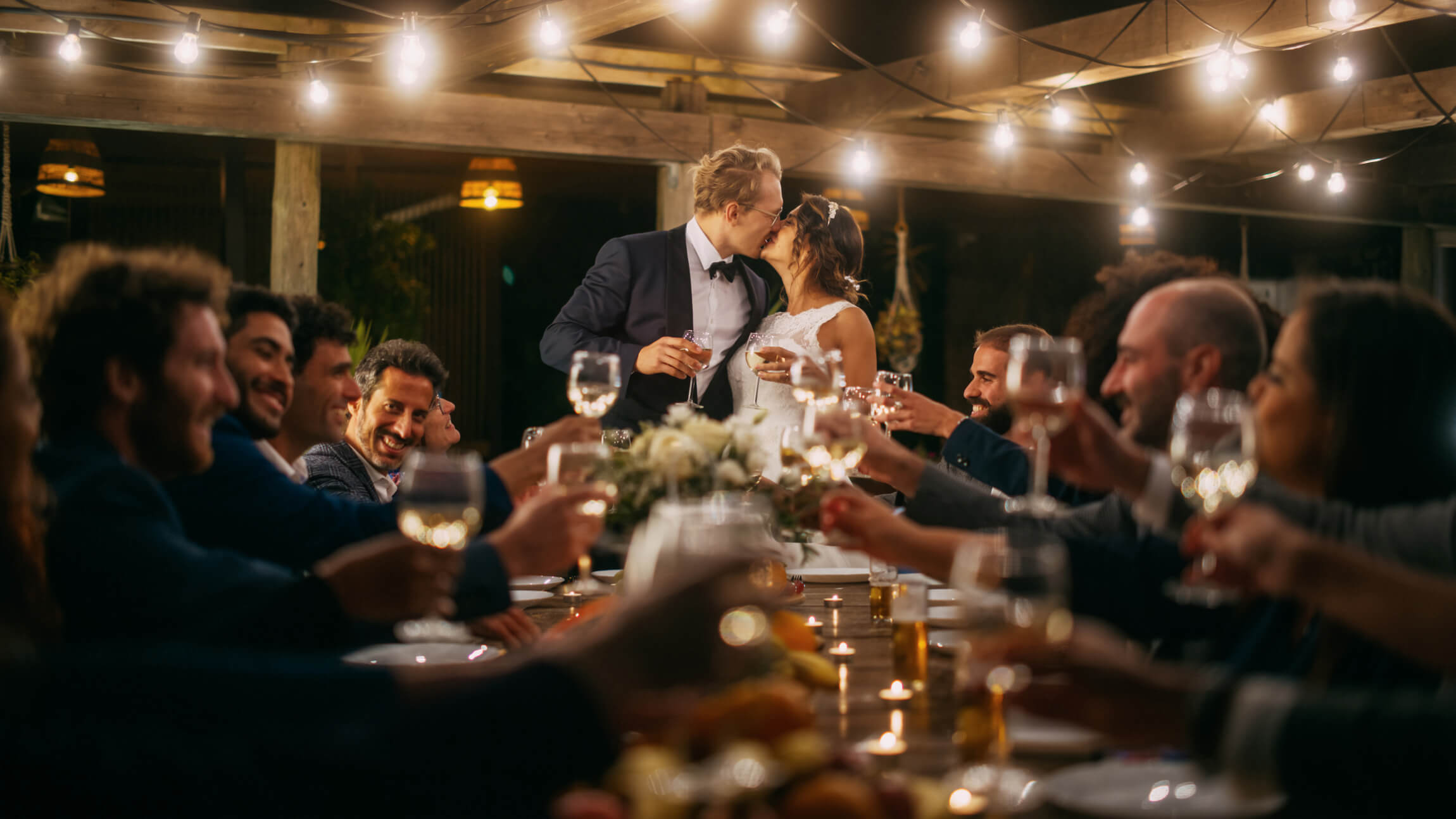 A bride and groom kiss at the head of a table while wedding guests cheers.