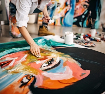 A painter uses their hands to paint a large, colorful picture of a woman.
