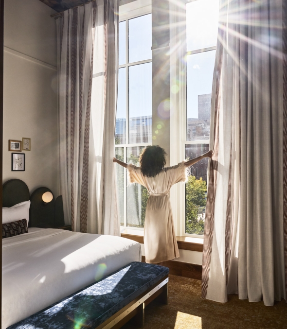 A woman opening her curtains to the sun shining in while staying at The Crawford Hotel